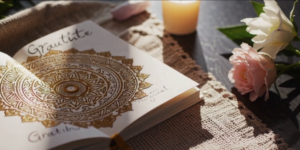 An open gratitude journal with a gold mandala design lies on a textured mat beside Matt Fraser—a lit candle—pink roses, and a crystal on a dark wooden surface.